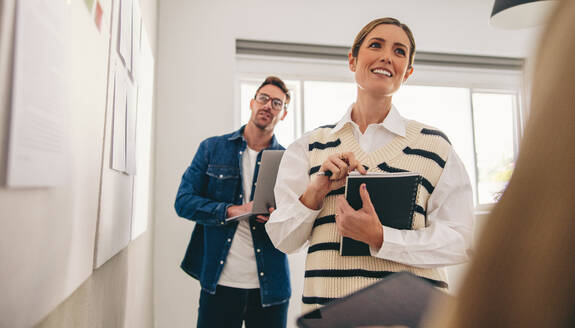 Business colleagues analysing some reports on a notice board during a meeting in a creative office. Group of cheerful businesspeople working as a team in a modern workplace. - JLPSF12922