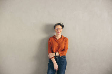 Mature businesswoman with eyeglasses smiling at the camera while standing against a grey wall. Happy businesswoman wearing business casual in a modern office. - JLPSF12925