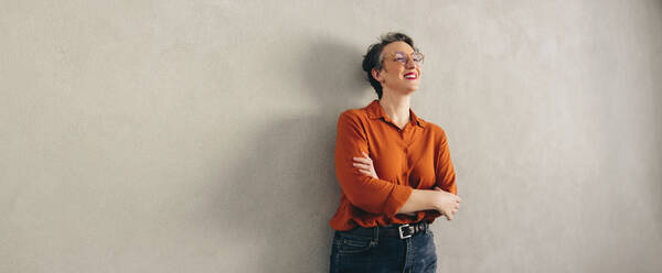 Cheerful businesswoman smiling happily while standing against a grey wall with her arms crossed. Mature businesswoman wearing business casual and eyeglasses in a modern office. - JLPSF12927