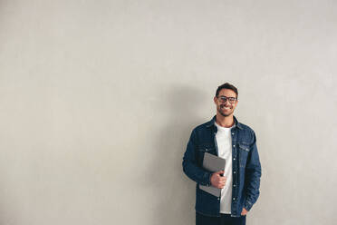 Fröhlicher junger Geschäftsmann, der in die Kamera lächelt, während er mit einem Laptop vor einer grauen Wand steht. Fröhlicher Geschäftsmann in Business Casual und mit Brille in einem modernen Büro. - JLPSF12929