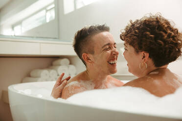 Unbeschwertes junges queeres Paar, das ein romantisches Schaumbad zu Hause nimmt. Glückliches junges Paar, das Spaß zusammen in der Badewanne hat. Junges LGBTQ+ Paar, das Zeit miteinander im Badezimmer verbringt. - JLPSF12942