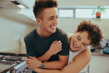 Junges queeres Paar lacht zusammen im Haus. Glückliches junges queeres Paar, das Spaß zusammen hat, während es in seiner Küche steht. Romantisches junges LGBTQ+ Paar, das sich liebevoll zu Hause verbindet. - JLPSF12945