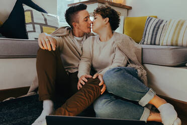 Süßes queeres Paar, das sich im Wohnzimmer küsst. Romantisches junges Paar, das sich küsst, während es zusammen im Wohnzimmer sitzt. Junges LGBTQ+ Paar, das einen intimen Moment miteinander teilt. - JLPSF12957