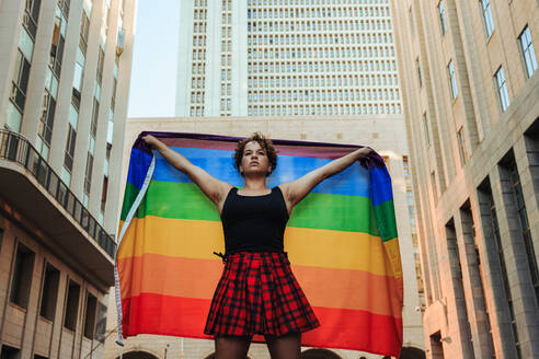 LGBTQ+ member flying the rainbow flag during a pride parade. Confident young lesbian woman celebrating gay pride during pride month. Young woman standing alone in the city during the day. - JLPSF12959