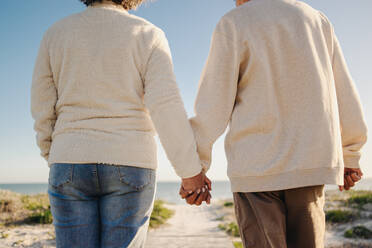 Senior couple holding hands while walking down a boardwalk at the beach. Unrecognisable elderly couple taking a refreshing seaside holiday after retirement. - JLPSF12964