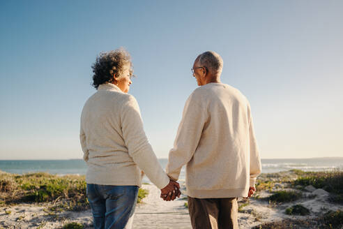 Rückansicht eines glücklichen älteren Paares, das sich an den Händen hält, während es an der Strandpromenade spazieren geht. Romantisches älteres Paar, das nach seiner Pensionierung einen erfrischenden Urlaub am Meer verbringt. - JLPSF12965