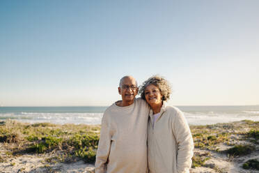 Porträt eines glücklichen älteren Paares, das gemeinsam am Strand steht und in die Kamera lächelt. Romantisches älteres Paar, das nach seiner Pensionierung einen entspannenden Urlaub am Meer verbringt. - JLPSF12967