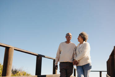 Romantic senior couple holding hands and smiling while taking a walk along a boardwalk. Affectionate elderly couple enjoying a refreshing beach holiday after retirement. - JLPSF12986