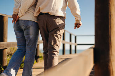 Rückansicht eines reifen Paares, das sich bei einem Spaziergang an der Strandpromenade an den Händen hält. Romantisches älteres Paar, das nach seiner Pensionierung einen erfrischenden Strandurlaub macht. - JLPSF12987
