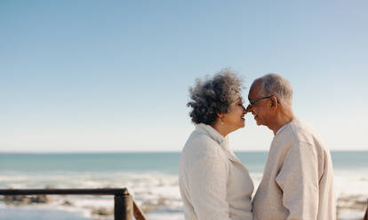 Being romantic by the ocean. Happy senior couple smiling and touching their noses together while standing on a seaside foot bridge. Retired elderly couple spending some quality time together. - JLPSF12992