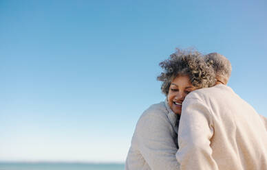 Zärtliche ältere Frau, die sich am Strand an die Schulter ihres Mannes lehnt und glücklich lächelt. Romantisches älteres Paar, das nach seiner Pensionierung einen erfrischenden Urlaub am Meer genießt. - JLPSF12994
