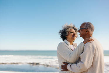 Romantic senior couple smiling happily while standing together at the beach. Cheerful elderly couple enjoying a refreshing seaside getaway after retirement. - JLPSF12995