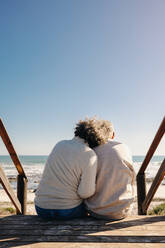 Back view of an elderly couple getting a refreshing view of the ocean while sitting on a wooden foot bridge. Retired senior couple spending some quality time together at the beach. - JLPSF12998