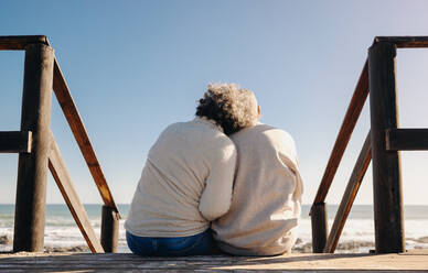 Rearview of a senior couple getting a refreshing view of the ocean while sitting on a wooden foot bridge. Retired elderly couple spending some quality time together by the ocean. - JLPSF12999