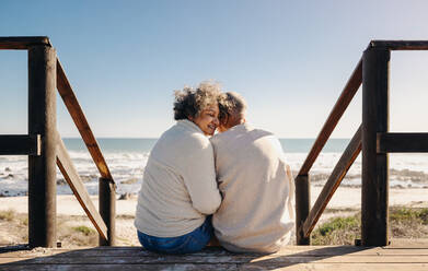 Romantic senior woman smiling happily while sitting on a wooden seaside bridge with her husband. Retired elderly couple spending some quality time together at the beach. - JLPSF13001
