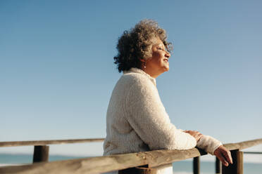 Glückliche ältere Frau genießt die Sonne am Strand, während sie auf der Promenade am Meer steht. Fröhliche ältere Frau, die nach ihrer Pensionierung einen erfrischenden Urlaub macht. - JLPSF13005