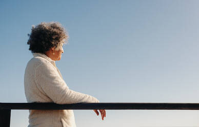 Eine ältere Frau im Ruhestand, die auf einer Strandpromenade steht und den Blick auf das Meer genießt. Eine glückliche ältere Frau, die nach ihrer Pensionierung einen erfrischenden Urlaub am Meer genießt. - JLPSF13008