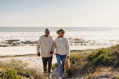 Fröhliches älteres Paar lächelt glücklich, während es nach einem Picknick vom Strand weggeht. Romantisches älteres Paar, das nach seiner Pensionierung einen Ausflug ans Meer genießt. - JLPSF13011