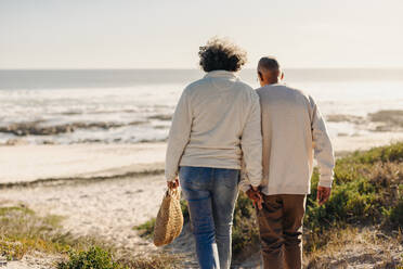 Rückansicht eines romantischen älteren Paares, das sich an den Händen hält, während es zum Strand geht, um dort ein Picknick zu machen. - JLPSF13012