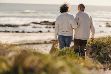 Rückenansicht eines romantischen älteren Paares, das zum Strand geht, um dort ein Picknick zu machen. Glückliches älteres Paar, das nach seiner Pensionierung Urlaub am Meer macht. - JLPSF13015