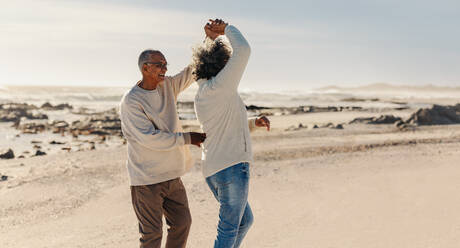 Romantic elderly couple dancing together by the beach. Happy mature couple having a good time next to the ocean. Cheerful senior couple enjoying their retirement days together. - JLPSF13020