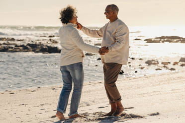 Happy elderly couple smiling and dancing together on beach sand. Mature couple having a good time next to the ocean. Cheerful senior couple enjoying their retirement days together. - JLPSF13021