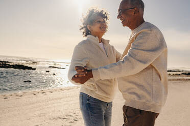 Cheerful senior couple smiling while dancing together at the beach. Happy elderly couple having a good time next to the ocean. Mature couple enjoying their retirement days together. - JLPSF13023