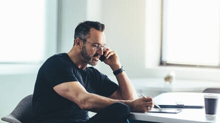 Unternehmer, der auf sein digitales Tablet schaut und mit seinem Handy spricht, während er an seinem Schreibtisch sitzt. Geschäftsmann, der im Büro arbeitet. - JLPSF13036