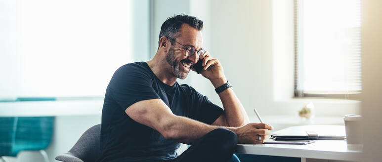 Lächelnder Mann bei der Arbeit, der mit seinem Handy telefoniert und an seinem digitalen Tablet arbeitet. Fröhlicher Geschäftsmann bei der Arbeit im Büro. - JLPSF13038