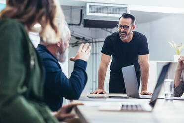 Glücklicher reifer Geschäftsmann im Gespräch mit Kollegen während einer Besprechung. Lächelnder männlicher Fachmann, der mit seinem Team neue Ideen diskutiert. - JLPSF13081