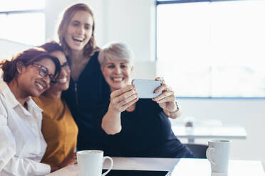 Lächelnde Geschäftsfrauen machen ein Selfie im Büro. Vier Geschäftsleute machen ein Selfie mit dem Handy in der Pause. - JLPSF13092