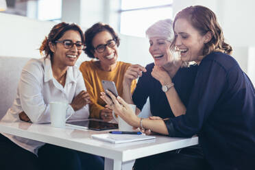 Glückliche Frauen zeigen ihr Telefon den Kollegen. Vier Geschäftsfrauen in der Pause im Büro schauen auf ihr Handy. - JLPSF13096