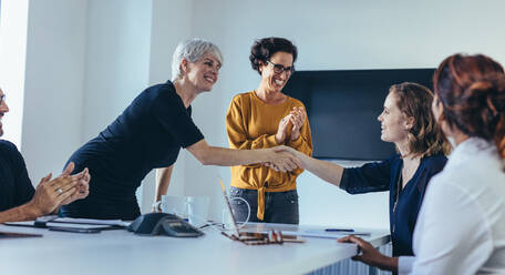 Geschäftsfrauen schütteln sich die Hände und lächeln während eines Geschäftstreffens mit einem Team, das in die Hände klatscht. Das Team gratuliert und würdigt die Leistung eines Kollegen im Besprechungsraum. - JLPSF13105