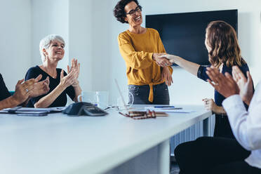 Businesswomen shaking hands and during a business meeting. Female professionals shaking hands with team sitting by clapping hands after a successful meeting. - JLPSF13108