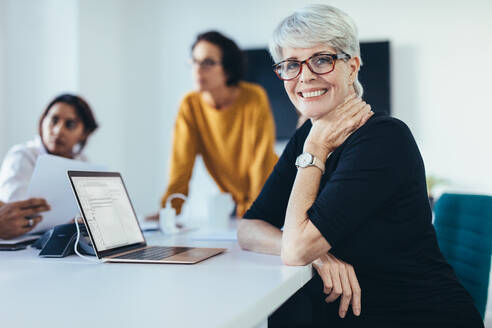 Successful mid adult woman sitting at a meeting with colleagues in boardroom. Female with coworkers in conference room looking at camera and smiling. - JLPSF13117