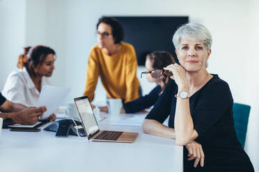 Reife Geschäftsfrau in einem Besprechungsraum mit Kollegen, die im Hintergrund diskutieren. Selbstbewusste weibliche Fachkraft in einer Vorstandssitzung. - JLPSF13118