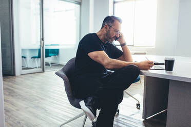 Männlicher Unternehmer, der auf sein digitales Tablet schaut und am Handy spricht, während er im Büro sitzt. Geschäftsmann, der im Büro arbeitet. - JLPSF13154
