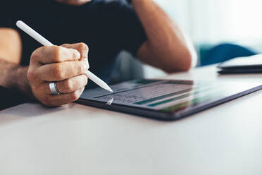 Nahaufnahme eines Geschäftsmannes, der mit einem digitalen Stift an einem Tablet arbeitet. Ein Mann sitzt am Tisch und arbeitet im Büro, der Fokus liegt auf dem Tablet-PC und dem digitalen Stift. - JLPSF13158