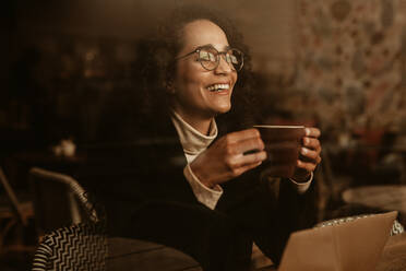 Frau mit Brille hält eine Kaffeetasse und lächelt im Café. Frau mit Kaffeetasse sitzt in einem Café. - JLPSF13160