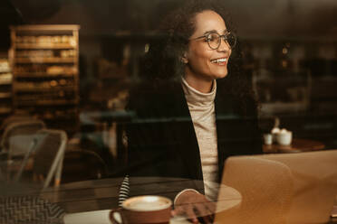 Geschäftsfrau in einem Café mit Laptop, die aus dem Fenster schaut und lächelt. Frau sitzt in einem Café mit ihrem Laptop. - JLPSF13163