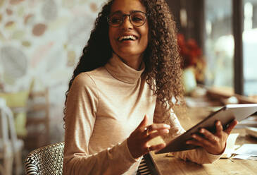 Lächelnde Frau, die in einem Café mit ihrem digitalen Tablet sitzt. Frau mit lockigem Haar, die wegschaut und lächelt, während sie an einem Kaffeetisch sitzt. - JLPSF13175