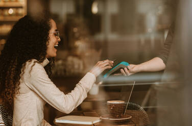 Smiling woman paying using NFC technology in a cafe to pay her bill. Female customer paying her bill using a smart phone and an electronic reader at coffee shop. - JLPSF13180