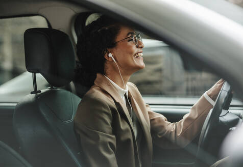 Geschäftsfrau mit Kopfhörern, die mit ihrem Auto ins Büro fährt und lächelt. Frau, die auf dem Weg zur Arbeit Musik hört. - JLPSF13185