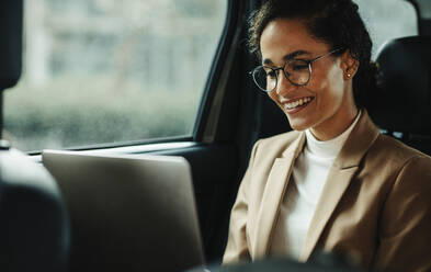 Lächelnde Geschäftsfrau, die mit einem Laptop arbeitet, während sie in einem Taxi fährt. Frau mit Laptop auf dem Rücksitz eines Autos. - JLPSF13187