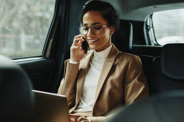 Geschäftsfrau, die an einem Laptop arbeitet und telefoniert, während sie in einem Taxi fährt. Frau sitzt auf dem Rücksitz eines Autos und benutzt Laptop und Telefon. - JLPSF13188