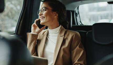 Businesswoman sitting on backseat of her car and talking on mobile phone. Businesswoman using phone while traveling by a car. - JLPSF13189