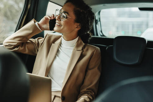 Fröhliche Frau im Geschäftsanzug auf dem Rücksitz ihres Autos sitzend und mit dem Handy telefonierend. Geschäftsfrau beim Telefonieren während der Fahrt im Auto. - JLPSF13190