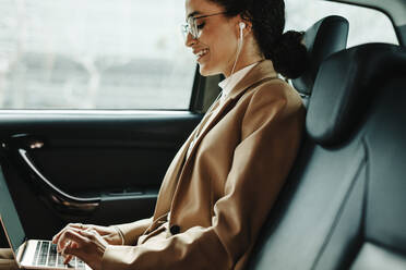 Seitenansicht einer lächelnden Geschäftsfrau, die während einer Taxifahrt an einem Laptop arbeitet. Frau benutzt Laptop auf dem Rücksitz eines Autos. - JLPSF13192
