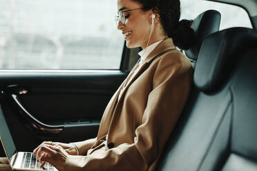 Side view of a smiling businesswoman working on laptop while travelling in a taxi. Woman using laptop in back seat of car. - JLPSF13192