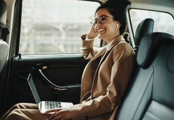 Lächelnde Geschäftsfrau, die mit einem Laptop in einem Taxi unterwegs ist. Frau mit Laptop, die auf dem Rücksitz ihres Autos sitzt, wegschaut und lächelt. - JLPSF13193